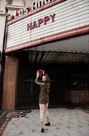 Young woman holding a heart-shaped box standing in front of the Los Angeles Theatre with the word 'Happy' on the marquee.