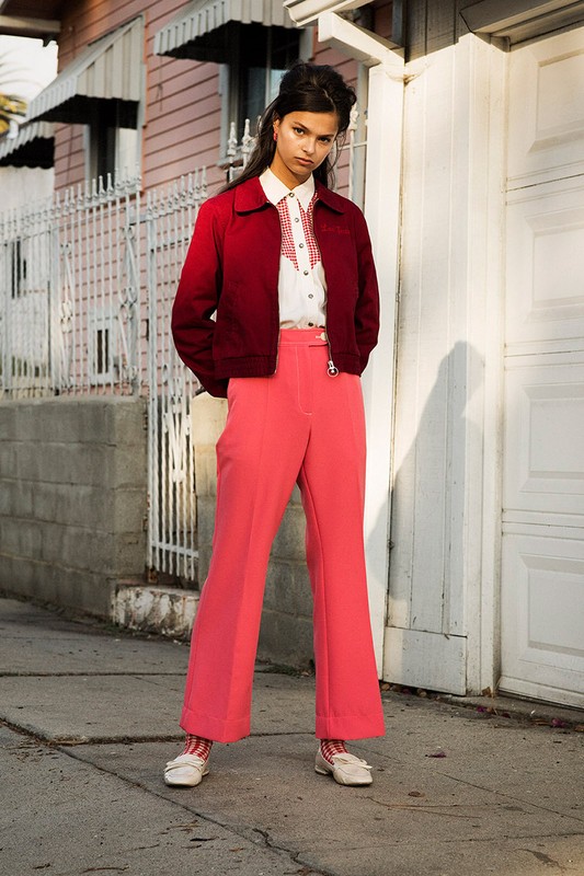 Young woman in a red jacket and coral pants stands on a sidewalk near a light pink house with a white fence during daytime.