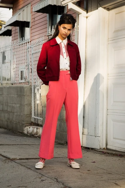 Young woman in a red jacket and coral pants stands on a sidewalk near a light pink house with a white fence during daytime.
