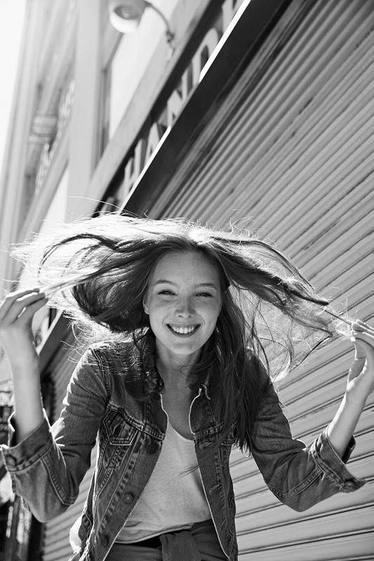 Black and white portrait of a smiling woman with long, flowing hair standing outside a building with a closed metal shutter.