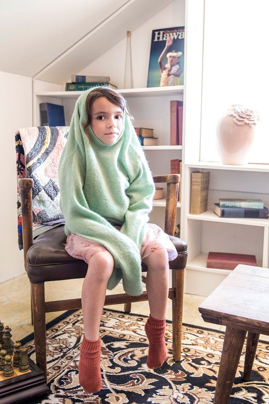 Young girl sits on a chair wearing a green sweater that is covering her head, with a bookshelf and Hawaiian magazine in background.