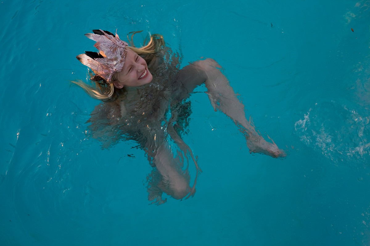 Woman floats in blue water with a feathered headpiece, smiling in an outdoor setting, creating a vibrant and lively visual.