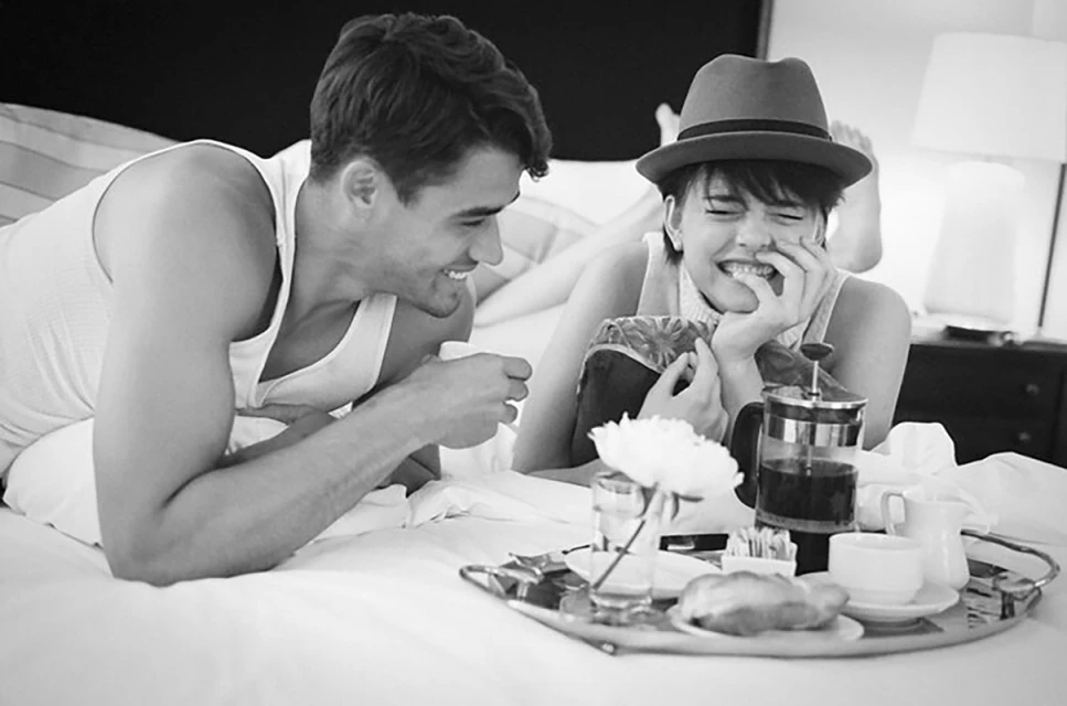 Black-and-white photo of a smiling couple lying on a bed with a breakfast tray featuring coffee, pastries, and a flower in a vase.