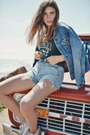 A woman in denim shorts and jacket sits on the hood of an orange truck against a blue sky. Rocks are visible in the background.