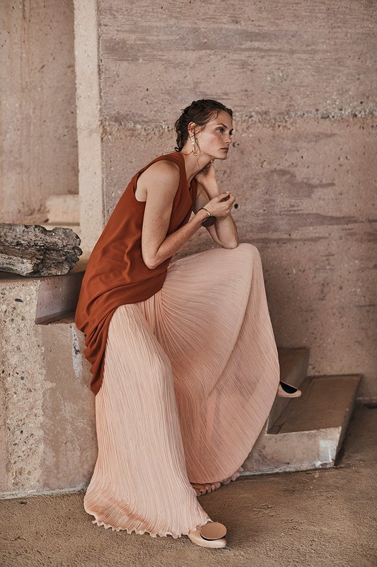 Elegant woman in rust-colored top and flowing peach skirt seated on concrete steps, complemented by minimalist jewelry and neutral flats.