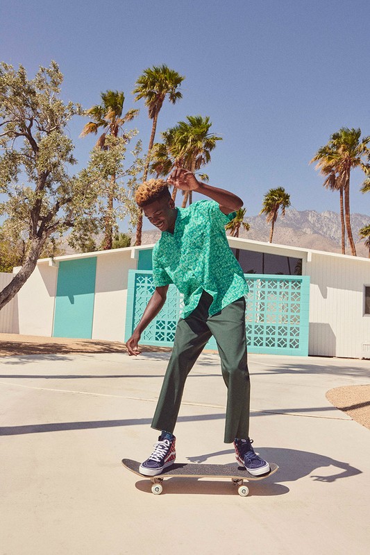 A young Black man skateboarding in front of a modern house with palm trees on a sunny day, wearing a green patterned shirt and dark trousers.