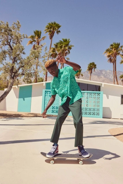 A young Black man skateboarding in front of a modern house with palm trees on a sunny day, wearing a green patterned shirt and dark trousers.