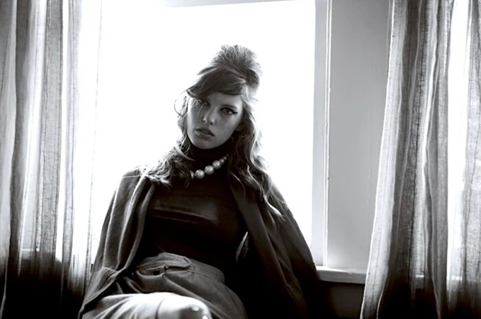 Black and white portrait of a stylish adult woman with a retro hairstyle, wearing a dark turtleneck, draped cape, and pearl necklace.