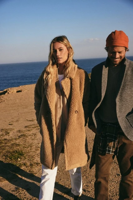 Outdoor fashion shot of a woman in a brown teddy coat and a man in a grey jacket, posing on a rocky cliff near the ocean under blue sky.