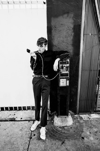 A young adult male in a black outfit stands near a vintage payphone on a city street in black and white, creating an editorial fashion image.