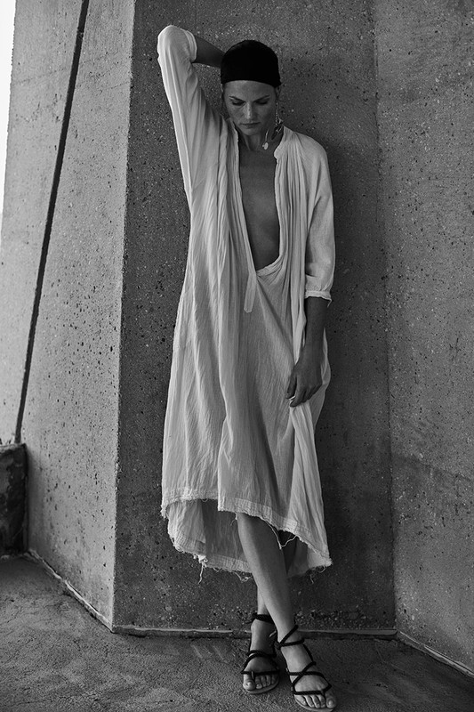 Monochrome portrait of an adult female wearing a light-colored dress and dark head covering, leaning against a textured concrete wall indoors.