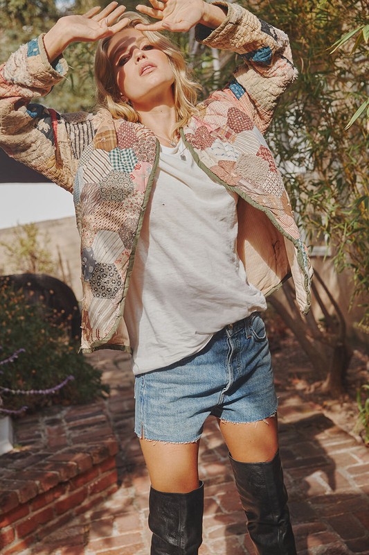 Woman in a patchwork jacket poses outside in denim shorts and high boots, hands raised to the sun, creating a summery, casual fashion shot.