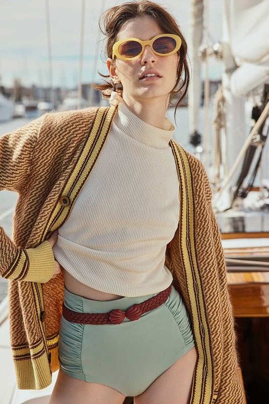 A woman poses on a boat in a brown cardigan, ivory top, and high-waisted swimsuit bottoms with yellow sunglasses and neutral lighting.