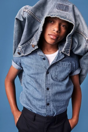 Young boy wearing a blue denim shirt with a denim jacket draped over his head against a blue background