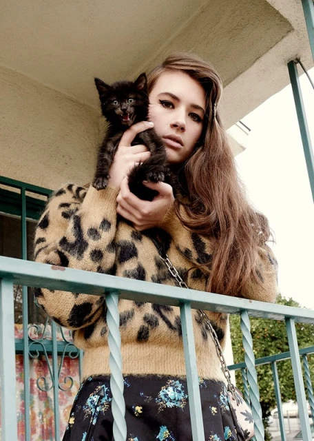 A young adult woman with long brown hair holds a small black kitten with its mouth open, wearing a leopard print sweater on a balcony.