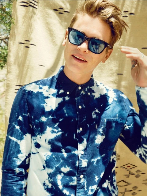 Portrait of a young man with blonde hair wearing sunglasses and a blue-and-white tie-dye shirt, posing against a patterned backdrop.