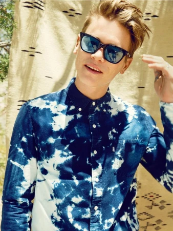 Portrait of a young man with blonde hair wearing sunglasses and a blue-and-white tie-dye shirt, posing against a patterned backdrop.