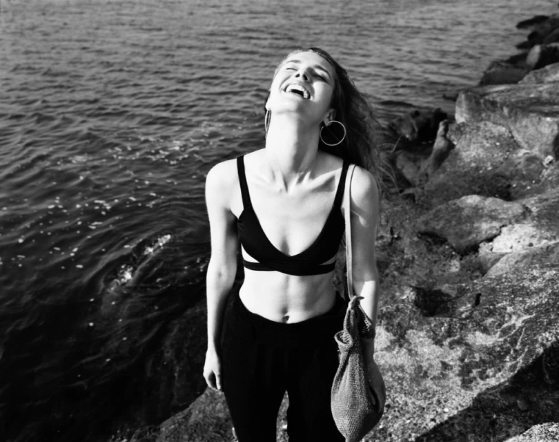 A young woman with long hair laughs joyously near a rocky shore with ocean waves, wearing a bra top and black pants, holding a mesh bag.