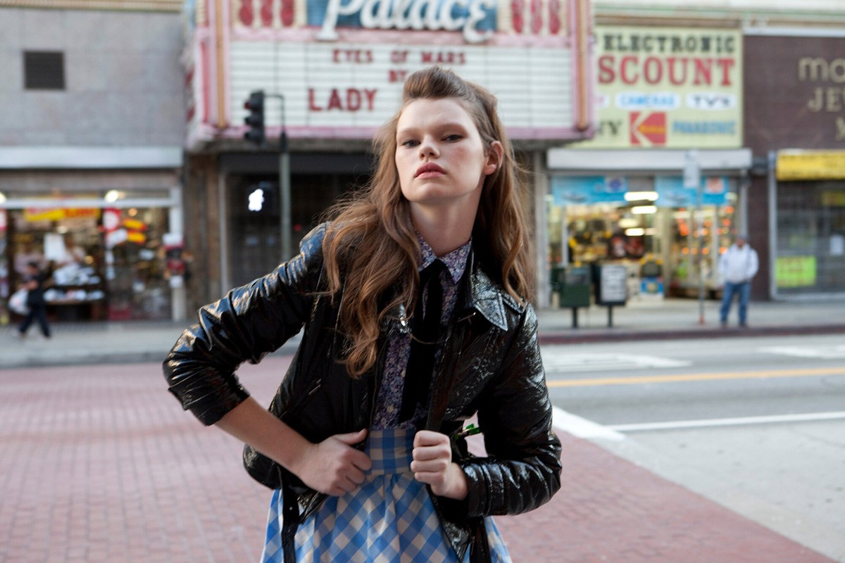 A young woman in a black jacket and plaid skirt stands on a city street with vintage storefronts in the background, creating a retro fashion vibe.