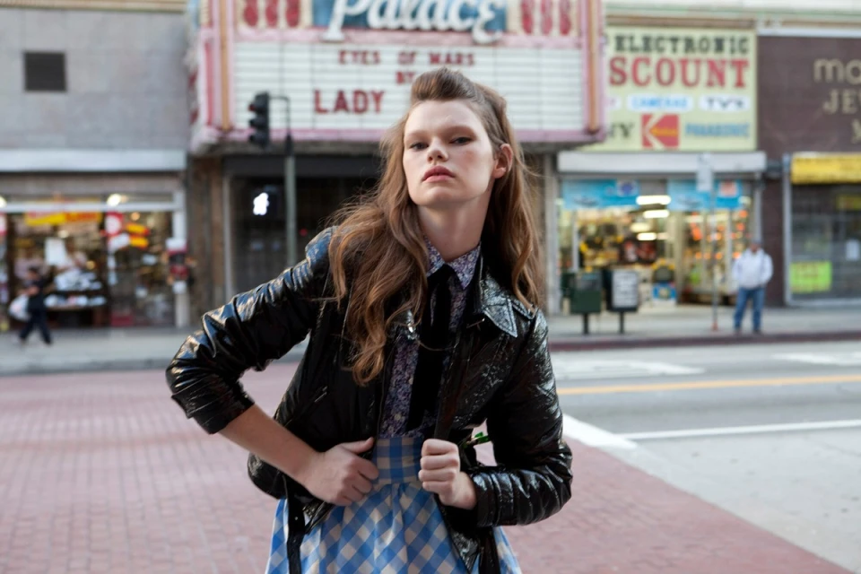 A young woman in a black jacket and plaid skirt stands on a city street with vintage storefronts in the background, creating a retro fashion vibe.