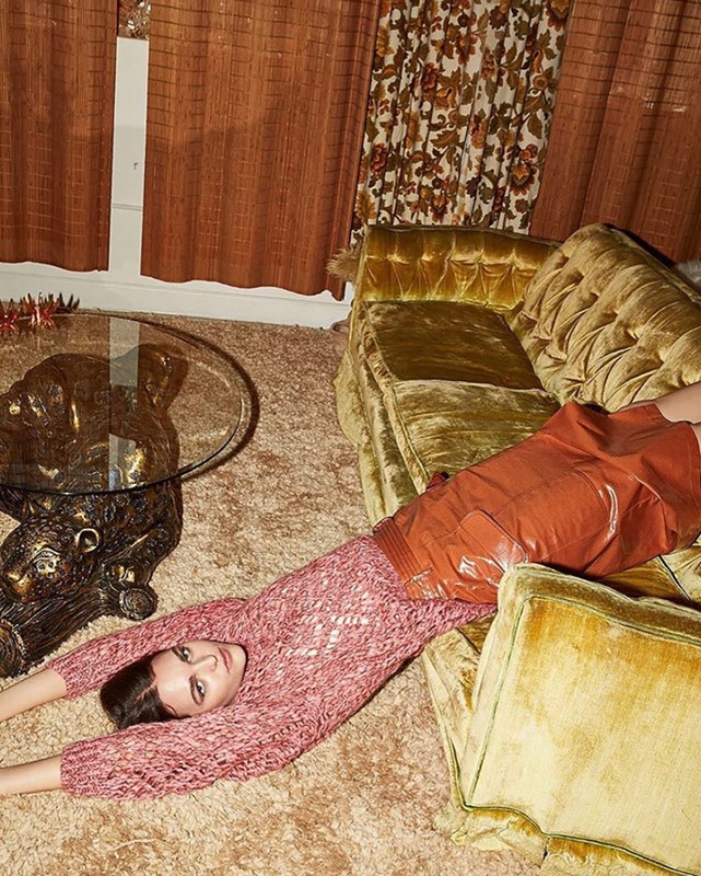 An adult woman lying on a shag carpet with a textured sweater and leather skirt, next to a glass table and vintage gold velvet couch.