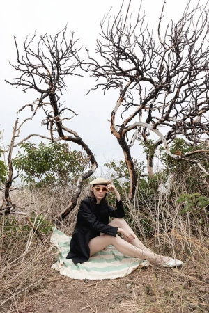 A seated woman wearing a black jacket and beret on a patterned blanket outdoors under bare trees styled by Steph Ashmore