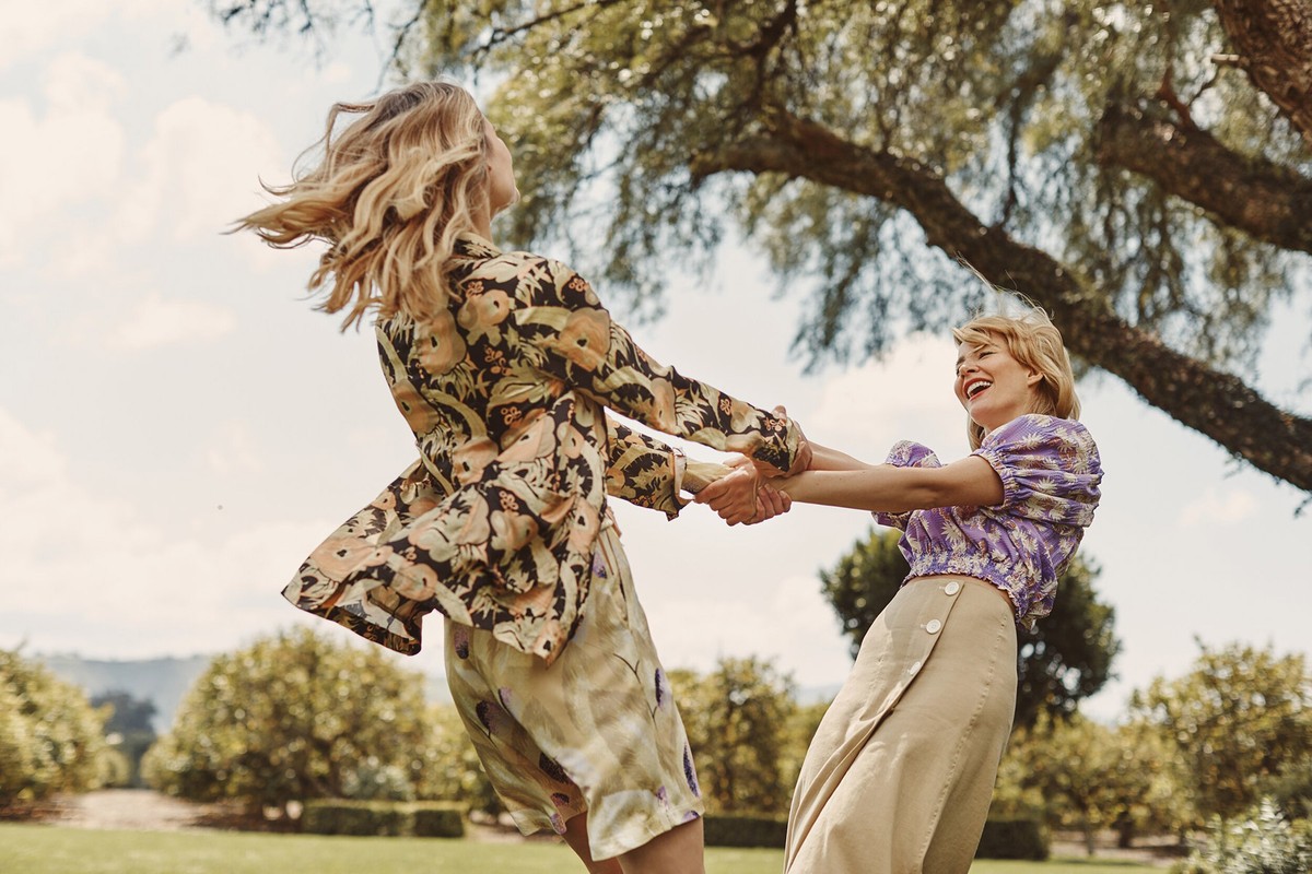 Two adult women in stylish floral print outfits joyfully spin in a circle on a grassy field under a large leafy tree on a sunny day.