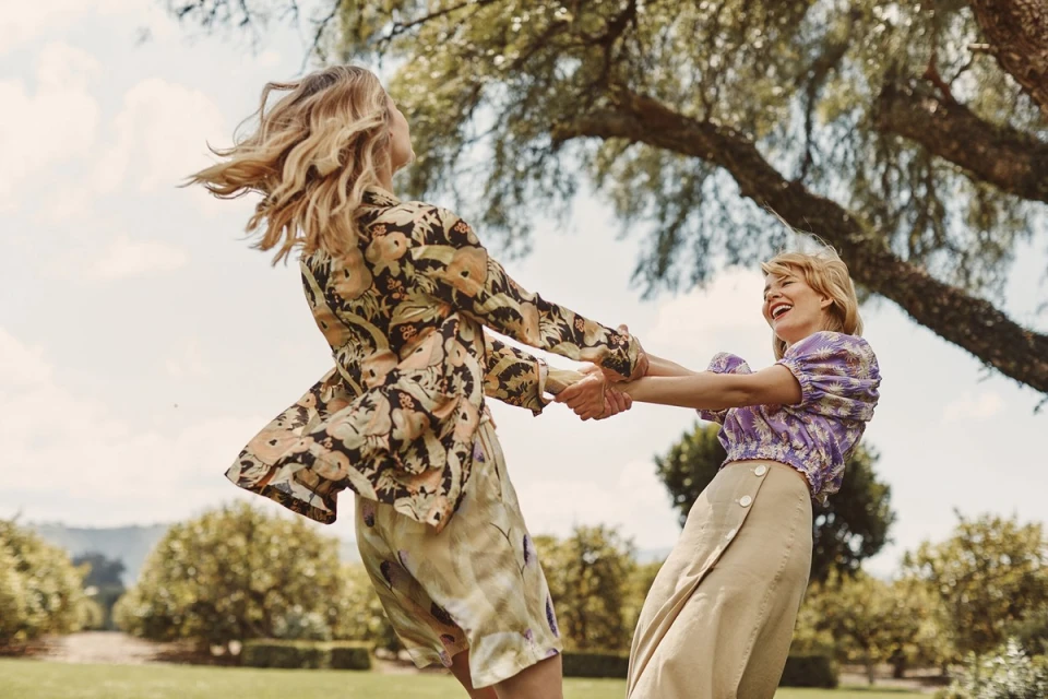 Two adult women in stylish floral print outfits joyfully spin in a circle on a grassy field under a large leafy tree on a sunny day.