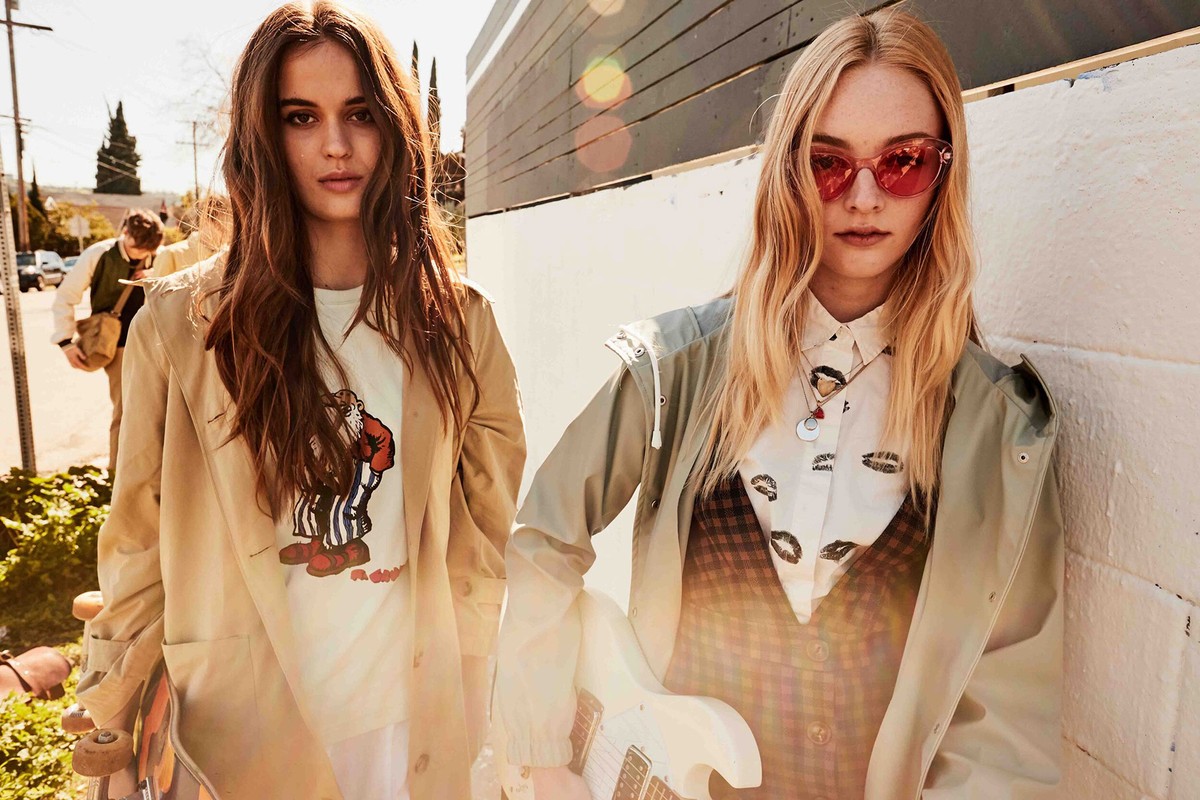 Two young women in casual fashion, one with sunglasses and a guitar, stand outdoors against a building on a sunny day, fashion editorial.
