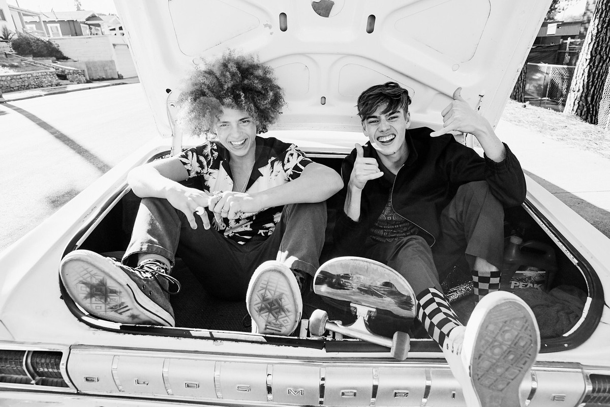 Two young men smile and pose in the open trunk of a classic white car on a sunny street with a skateboard and patterned socks visible.