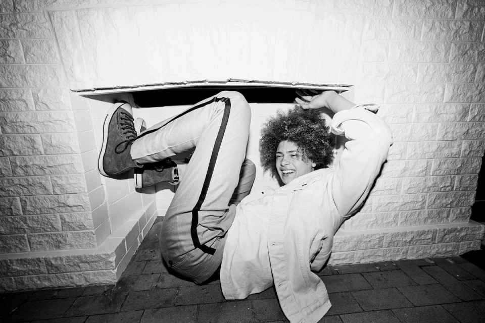 A person with afro hairstyle smiles in a black and white photo, wearing jeans and a shirt, and lying underneath a structure.