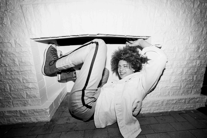 A person with afro hairstyle smiles in a black and white photo, wearing jeans and a shirt, and lying underneath a structure.