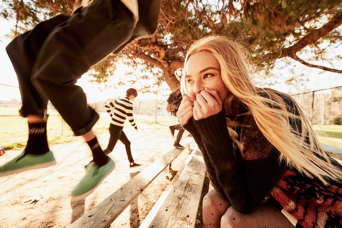 Young woman smiling by a park bench, surrounded by friends on a sunny day with trees in the background, and one friend leaping over the bench.
