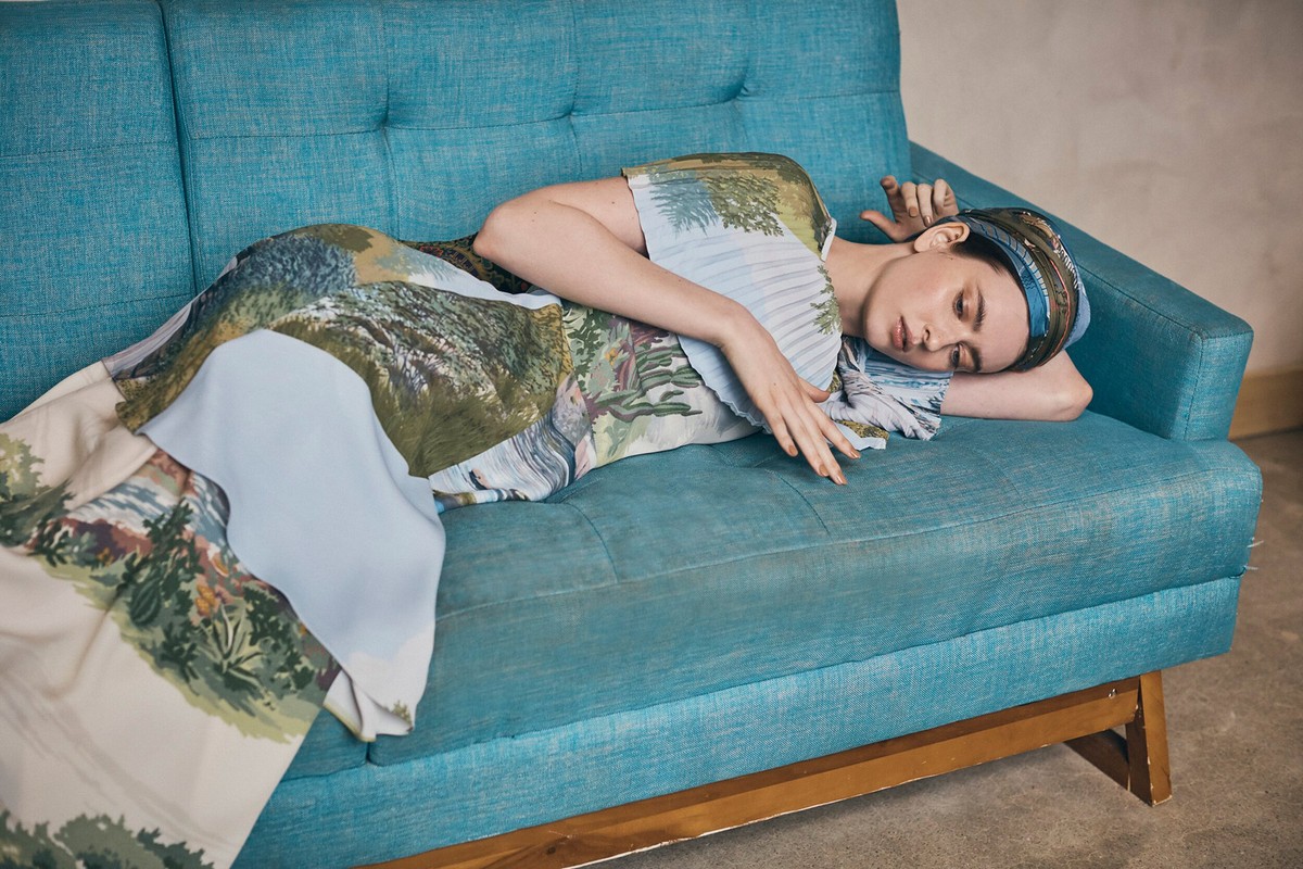 Adult woman with eyes closed in landscape-printed dress reclines on a blue couch indoors with a neutral wall background, shot at eye level.