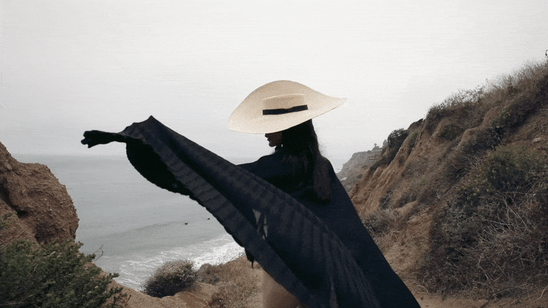 Woman wearing a wide-brim straw hat and a flowing black cape overlooking a hazy coastal ocean view