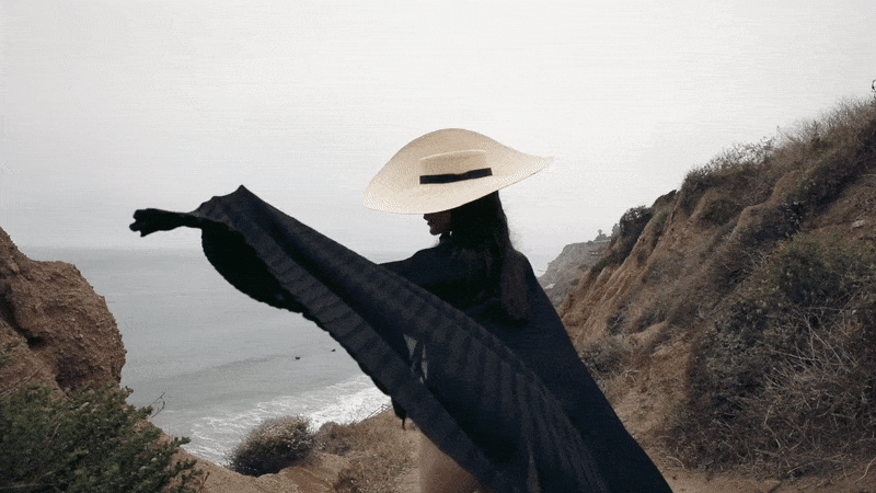 Woman wearing a wide-brim straw hat and a flowing black cape overlooking a hazy coastal ocean view