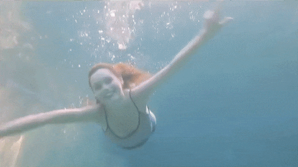 Underwater shot of a smiling adult woman swimming in clear blue water with her arms outstretched. Water bubbles are visible.
