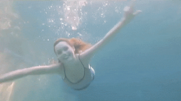 Underwater shot of a smiling adult woman swimming in clear blue water with her arms outstretched. Water bubbles are visible.