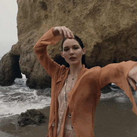 Woman in an orange shirt standing on a beach with rock formations in the background, posing with her arms and hands in the air.