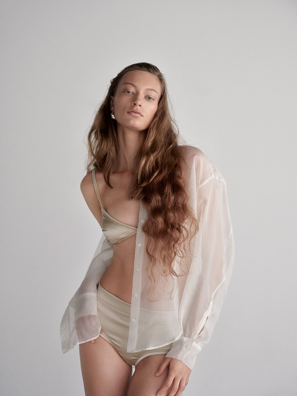 Woman with long wavy brown hair wearing a satin bralette, high-waisted briefs, and a sheer white button-down shirt against a neutral backdrop