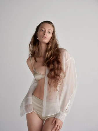Woman with long wavy brown hair wearing a satin bralette, high-waisted briefs, and a sheer white button-down shirt against a neutral backdrop