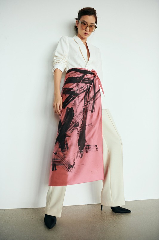 Woman in a white blazer and pink sarong-style skirt with black brushstroke design poses against a white wall in a studio setting.