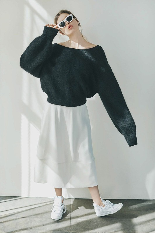 A young adult woman stands in a photo studio wearing a black sweater, white skirt, white sneakers, and sunglasses looking at the camera.