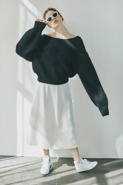 A young adult woman stands in a photo studio wearing a black sweater, white skirt, white sneakers, and sunglasses looking at the camera.