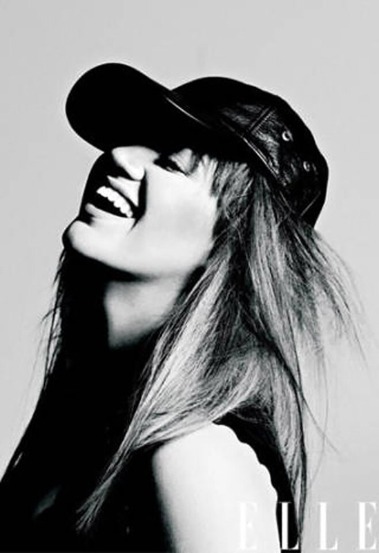 Monochrome headshot of a smiling woman wearing a leather baseball cap looking upward, contrasted against a light grey backdrop, and featuring the Elle logo.