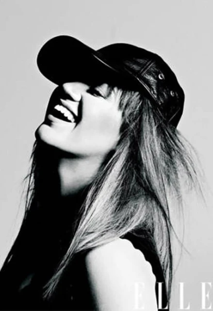 Monochrome headshot of a smiling woman wearing a leather baseball cap looking upward, contrasted against a light grey backdrop, and featuring the Elle logo.