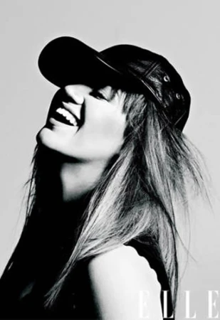 Monochrome headshot of a smiling woman wearing a leather baseball cap looking upward, contrasted against a light grey backdrop, and featuring the Elle logo.