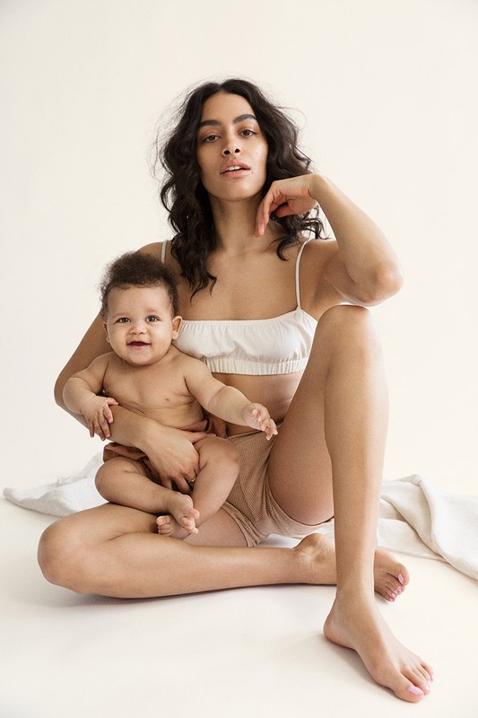 A light-skinned adult woman in bralette holds a baby on her lap in a portrait on an off-white seamless backdrop in soft lighting.