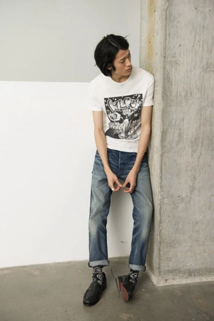 A lean young man leans against a corner wearing a graphic tee, denim jeans, and chunky leather shoes in a bright, casual setting.