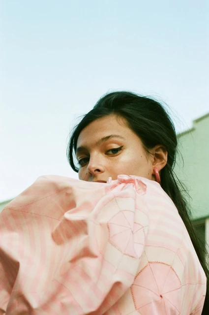 Close-up portrait of a brunette woman in a pink dress with umbrella pattern against a light blue and green exterior background.